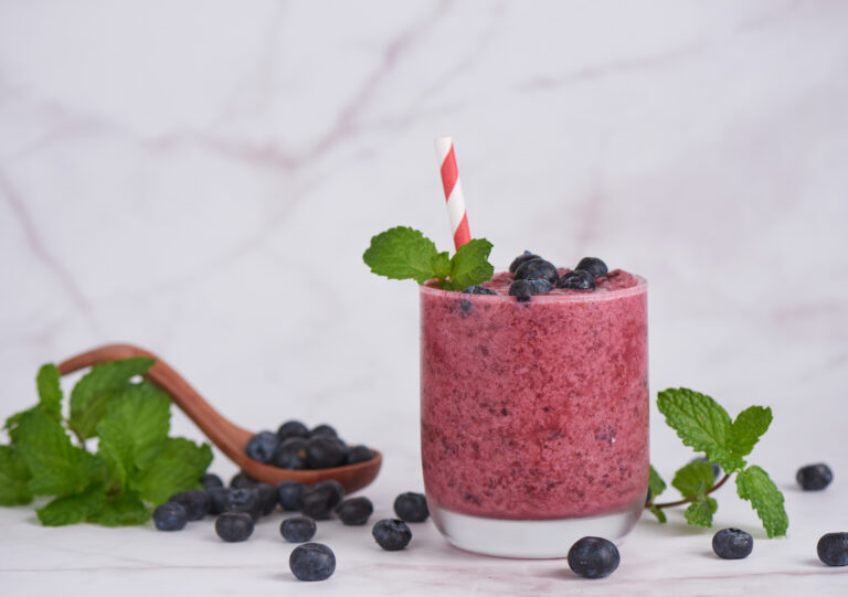 small glass full with rich red blue smoothie topped with blueberries with blueberries sprinkled around on a white background